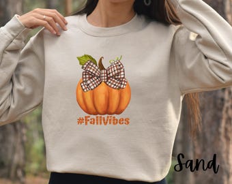 May include: A beige sweatshirt featuring a graphic of a pumpkin with a plaid bow and the text "#FallVibes".