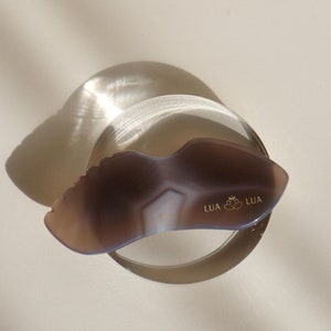 Puede incluir: Una herramienta de gua sha gris de forma irregular con una superficie lisa y pulida. La herramienta tiene un borde elevado y texturizado y las palabras "LUA LUA" están impresas en la parte inferior.