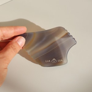 Puede incluir: Una herramienta de gua sha de ágata gris y marrón con un borde festoneado. La herramienta tiene la palabra "LUA" y un logotipo de flor de loto grabado.