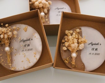 May include: Two small, round, white and brown resin keepsake boxes with dried flowers and gold glitter. The boxes are in brown cardboard boxes with the text "Ayssiah's 18th 19.11.2022" on the front.