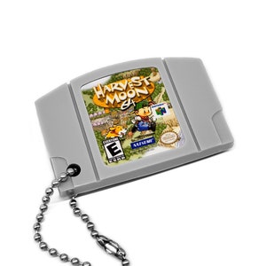 May include: A gray plastic keychain shaped like a Nintendo 64 game cartridge. The cartridge has a label with the text "Harvest Moon 64" and a cartoon image of a farmer. The keychain has a silver metal chain attached.