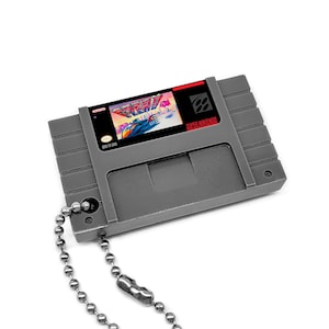 May include: A gray Super Nintendo Entertainment System (SNES) game cartridge keychain with a silver chain. The cartridge label features a digital render of the game "Aero the Acro-Bat 2".