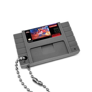 May include: A gray Super Nintendo cartridge keychain with a silver chain. The cartridge has a label with a digital render of the game Street Fighter Alpha 2.