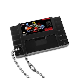 May include: Black Super Nintendo cartridge keychain with a silver chain. The cartridge label features a red and black graphic of a character from the game "Contra: Hard Corps".