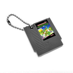 May include: A grey plastic keychain shaped like a Nintendo Entertainment System game cartridge. The cartridge has a label with the text "Teenage Mutant Ninja Turtles" and an image of the four turtles.