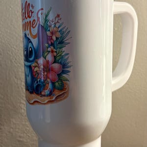 Może przedstawiać: Biały kubek podr&oacute;żny z uchwytem, z kolorową ilustracją Stitcha z Lilo & Stitch z tropikalnymi kwiatami i napisem "Hello Summer". Kubek ma przezroczystą pokrywę.