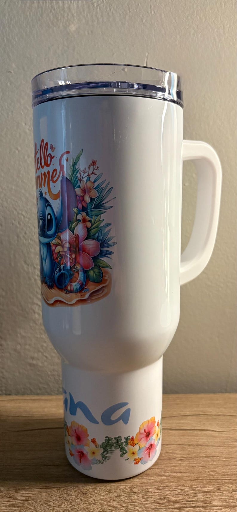 Może przedstawiać: Biały kubek podr&oacute;żny z uchwytem, z kolorową ilustracją Stitcha z Lilo & Stitch z tropikalnymi kwiatami i napisem "Hello Summer". Kubek ma przezroczystą pokrywę.