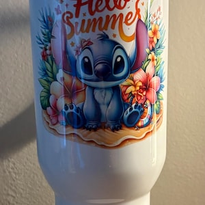 Może przedstawiać: Biały kubek z motywem Stitch z Lilo & Stitch, tropikalnymi kwiatami i napisem "Hello Summer". Na dole kubka znajduje się napis "hana" w niebieskim kolorze, z kwiatowymi detalami. Zabawny, letni artykuł do picia.