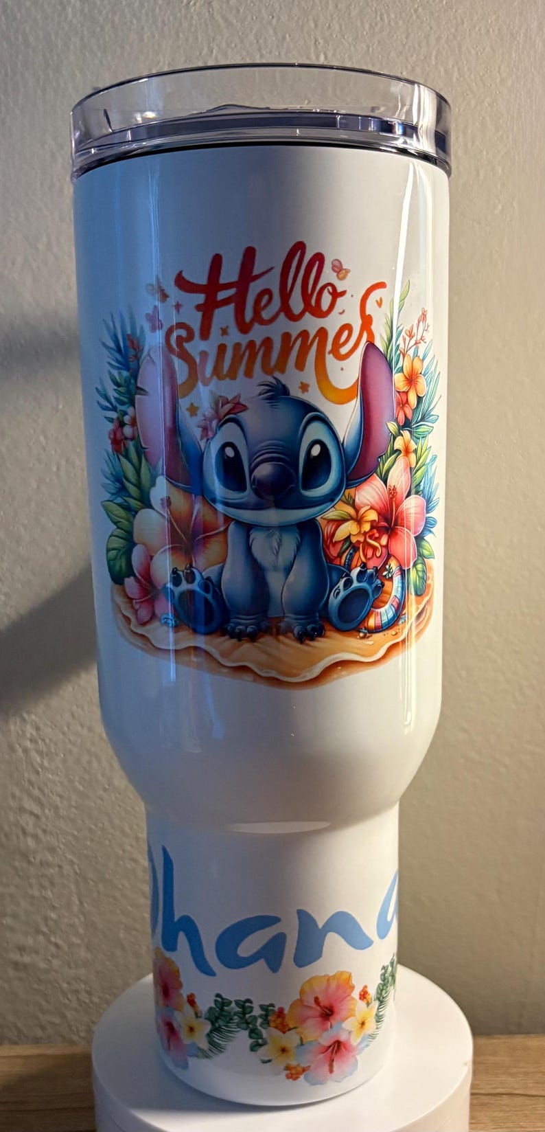 Może przedstawiać: Biały kubek z motywem Stitch z Lilo & Stitch, tropikalnymi kwiatami i napisem "Hello Summer". Na dole kubka znajduje się napis "hana" w niebieskim kolorze, z kwiatowymi detalami. Zabawny, letni artykuł do picia.