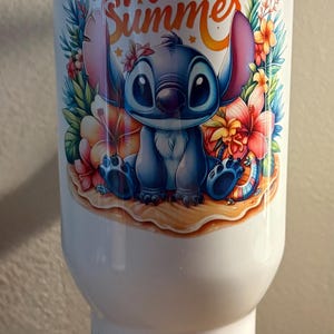 Może przedstawiać: Biały kubek z postacią Stitcha z Lilo & Stitch z napisem "Hello Summer". Projekt zawiera kolorowe kwiaty i scenę na plaży. D&oacute;ł kubka ma kwiatowy wz&oacute;r i imię "hana" w kolorze niebieskim.