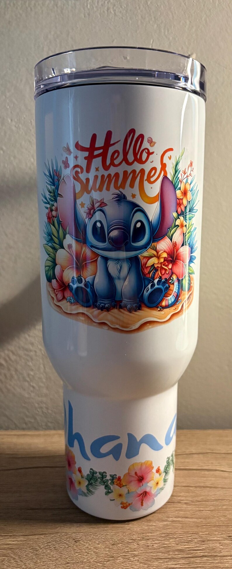 Może przedstawiać: Biały kubek z postacią Stitcha z Lilo & Stitch z napisem "Hello Summer". Projekt zawiera kolorowe kwiaty i scenę na plaży. D&oacute;ł kubka ma kwiatowy wz&oacute;r i imię "hana" w kolorze niebieskim.