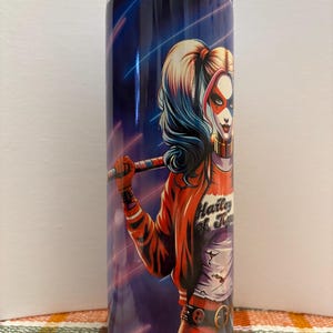 May include: A stainless steel tumbler featuring a vibrant illustration of a character with red, white, and blue hair and makeup, holding a baseball bat. The character is wearing a red jacket with the text "Harley & Ivy". The background is a dark blue with neon lines.