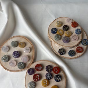 May include: A collection of small, round, polished stones in various colors including red, yellow, blue, purple, and grey, each engraved with a unique silver or gold symbol. These stones are arranged on three circular wooden slices with bark edges.