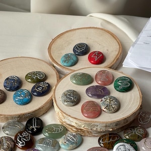 Peut inclure: Une collection de pierres ovales, lisses et polies, de différentes couleurs dont le bleu, le vert, le rouge, le violet et le blanc. Chaque pierre présente un symbole ou une icône gravé à la main, rempli de peinture argentée ou dorée.