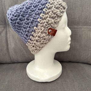 May include: A two-toned crochet beanie featuring a periwinkle blue upper section and a beige lower brim. The hat is adorned with a small, rectangular brown leather tag attached by a silver metal rivet, which is embossed with the text hand made with love.