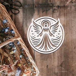 Puede incluir: Corte de madera blanco de un ángel con las alas extendidas. El ángel tiene un halo y está rodeado de estrellas. El corte está sobre un fondo de madera.