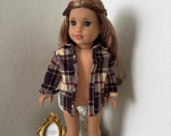 May include: A doll with long brown hair and freckles wearing an unbuttoned long-sleeved shirt featuring a brown, cream, and red plaid pattern. The doll has brown eyes and a neutral expression. Beside the doll is a small gold ornate picture frame.