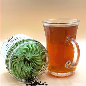 May include: A glass jar filled with a green and black whipped body scrub with the text "Mint Tea Body Scrub" on the label. A glass mug filled with brown tea sits next to the jar. Loose tea leaves are scattered on the surface.