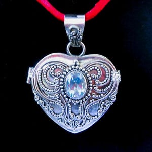 May include: A heart-shaped silver locket pendant featuring intricate beaded scrollwork and a central oval clear gemstone. The locket is suspended from a bright red cord, showcasing a detailed metal bail with decorative swirling patterns at the top.