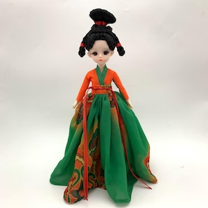 May include: A doll dressed in a traditional Asian-style outfit. The doll has black hair styled in an updo with red accents. The outfit features an orange top, a green skirt with an orange and brown pattern, and red ribbons.