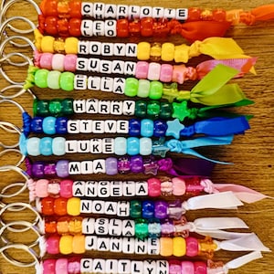 May include: A set of 12 colourful beaded keychains with names spelled out in white letter beads. Each keychain has a different colour scheme and a ribbon attached.