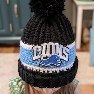 May include: A black knit beanie with a blue and white stripe and a pom pom on top. The beanie has a white and blue embroidered patch that says "LIONS" with a blue lion logo.