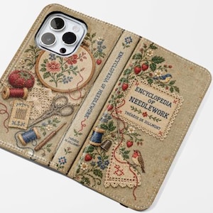 Peut inclure: Un étui de téléphone portefeuille beige avec un motif imprimé représentant une couverture de livre Encyclopedia of Needlework, des tambours à broder, des bobines de fil, des ciseaux et des motifs floraux, incluant une découpe pour appareil photo.
