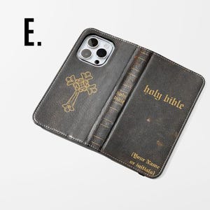 May include: A dark gray leather phone case with a gold cross and the words "holy bible" printed on it. The case is designed to look like a book, with the spine visible. The text "[Your Name or Initials]" is also printed on the case.