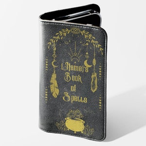 May include: A black leather phone case with a gold design. The design features a cauldron, feathers, and the text "The Namel's Book of Spells".
