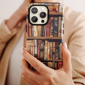 May include: A smartphone case featuring a detailed illustration of a bookshelf filled with various books. The case has a black border around the camera and a beige background. The books are in various colors, including red, brown, and beige.