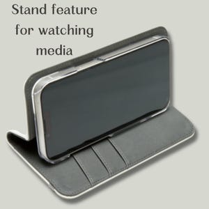 May include: A gray phone case with a stand feature for watching media. The case has a white border and a gray interior with card slots. The phone is in a clear case.