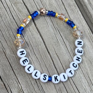 May include: A beaded bracelet with the word "HELLSKITCHEN" spelled out in white letter beads. The bracelet features blue, yellow, and clear beads, with a gold-colored clasp. The bracelet is displayed on a wooden surface.