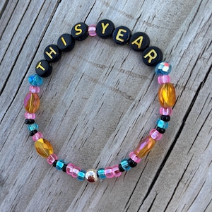 May include: A beaded bracelet with the words "THIS YEAR" spelled out in black letter beads with gold lettering. The bracelet features pink, blue, and gold beads, and a silver bead.