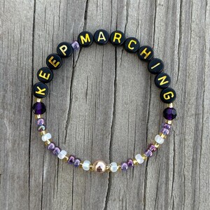 May include: A beaded bracelet with the words "KEEP MARCHING" spelled out in black and gold letter beads. The bracelet features purple, gold, and white beads, and a central gold-colored bead. The bracelet is set against a wooden background.