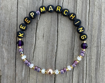 Op de afbeelding: Een kralenarmband met de woorden "KEEP MARCHING" in zwarte en gouden letterkralen. De armband heeft paarse, gouden en witte kralen en een centrale goudkleurige kraal. De armband ligt op een houten achtergrond.