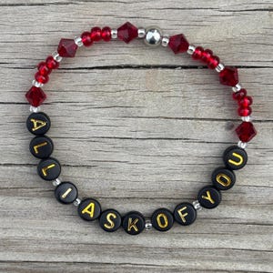 May include: A red and black beaded bracelet with the words "All I ask of you" spelled out in gold letters.