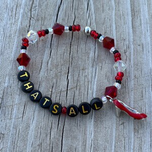 May include: A beaded bracelet featuring red and clear faceted beads, small black and red seed beads, and black round beads with the gold text "THATS ALL" printed on them. A red high-heeled shoe charm hangs from the bracelet, which is strung on elastic.