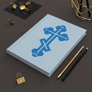 Könnte beinhalten: Ein hellblaues Notizbuch mit einem blauen Kreuz und den Buchstaben IC XC NI KA darauf. Das Notizbuch liegt auf einer braunen Lederoberfläche mit goldenen Büroklammern und Bleistiften.