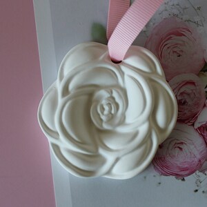 May include: A white ceramic rose-shaped air freshener with a pink ribbon attached. The air freshener is on a white background with a picture of pink flowers.