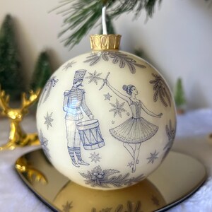 May include: Cream-coloured Christmas bauble with blue illustrations of a drummer and a ballerina. The bauble has a gold-coloured top and sits on a gold-coloured, heart-shaped plate. The background includes a Christmas tree and a gold reindeer.