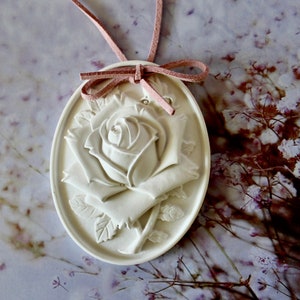 May include: A white ceramic rose pendant with a pink suede cord. The rose is detailed with a raised design and is oval shaped.