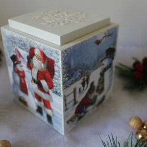May include: A white wooden box with a hinged lid decorated with a winter scene featuring Santa Claus, a snowman, and a penguin. The box has a white lid with a snowflake design.