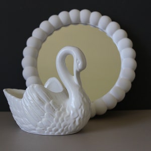 May include: White ceramic swan figurine with a decorative, round mirror behind it. The mirror has a white, beaded frame.