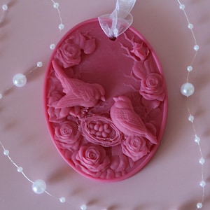 May include: A pink oval-shaped ceramic ornament with a raised design of two birds and a nest with eggs. The ornament is surrounded by a string of white pearls.