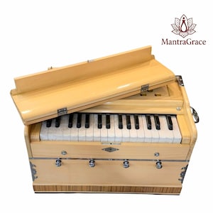 May include: A light-colored wooden harmonium musical instrument with a keyboard featuring white and black keys, a hinged lid, and four metal knobs on the front panel. The logo MantraGrace is visible in the upper right corner of the image.