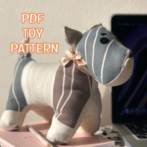 Könnte beinhalten: Ein handgefertigtes Stoffhundespielzeug mit einem gestreiften Muster in Blau-, Braun- und Weißtönen. Das Spielzeug hat eine kleine rosa Schleife und den Text "PDF TOY PATTERN" in Orange. Das Spielzeug liegt auf einem rosa Buch.