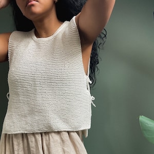 Può includere: Un top bianco in crochet dalla vestibilità ampia con un dettaglio di laccio sul lato. Il top è abbinato a una gonna in lino beige.