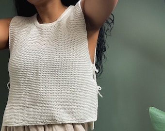May include: A white crochet tank top with a loose fit and a tie detail at the side. The tank top is paired with a beige linen skirt.
