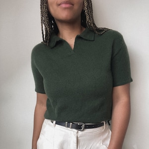 May include: A dark green short-sleeved knitted polo shirt with a collared neckline. The shirt is worn with a white pair of trousers and a black belt.