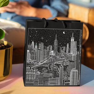 May include: A black gift bag featuring a detailed white illustration of the New York City skyline, including the Brooklyn Bridge, skyscrapers, and a starry night sky. The bag has black rope handles.